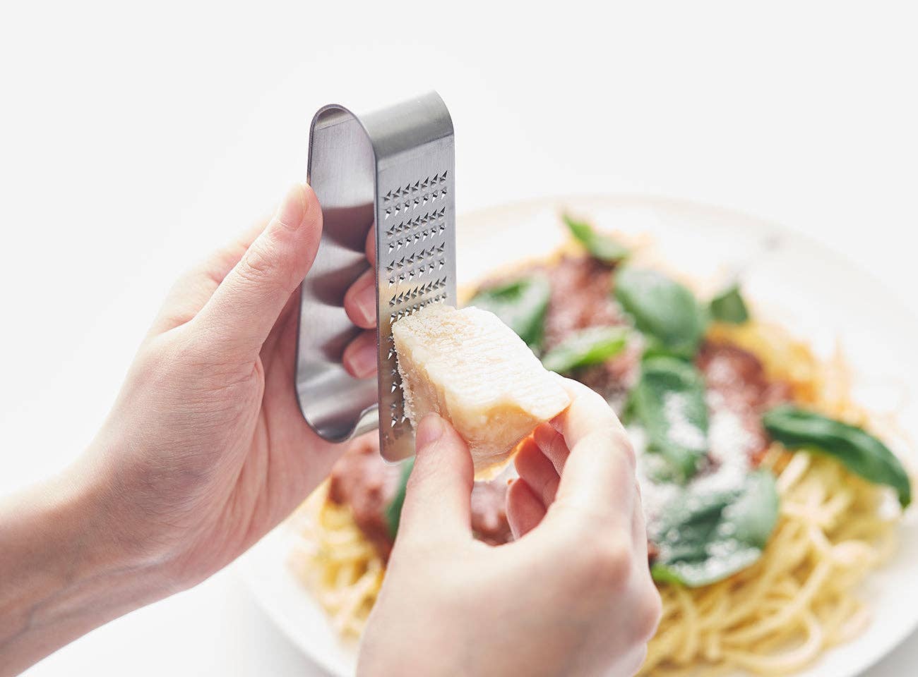 Hand holding a metal grater over a plate of pasta with greens and cheese.