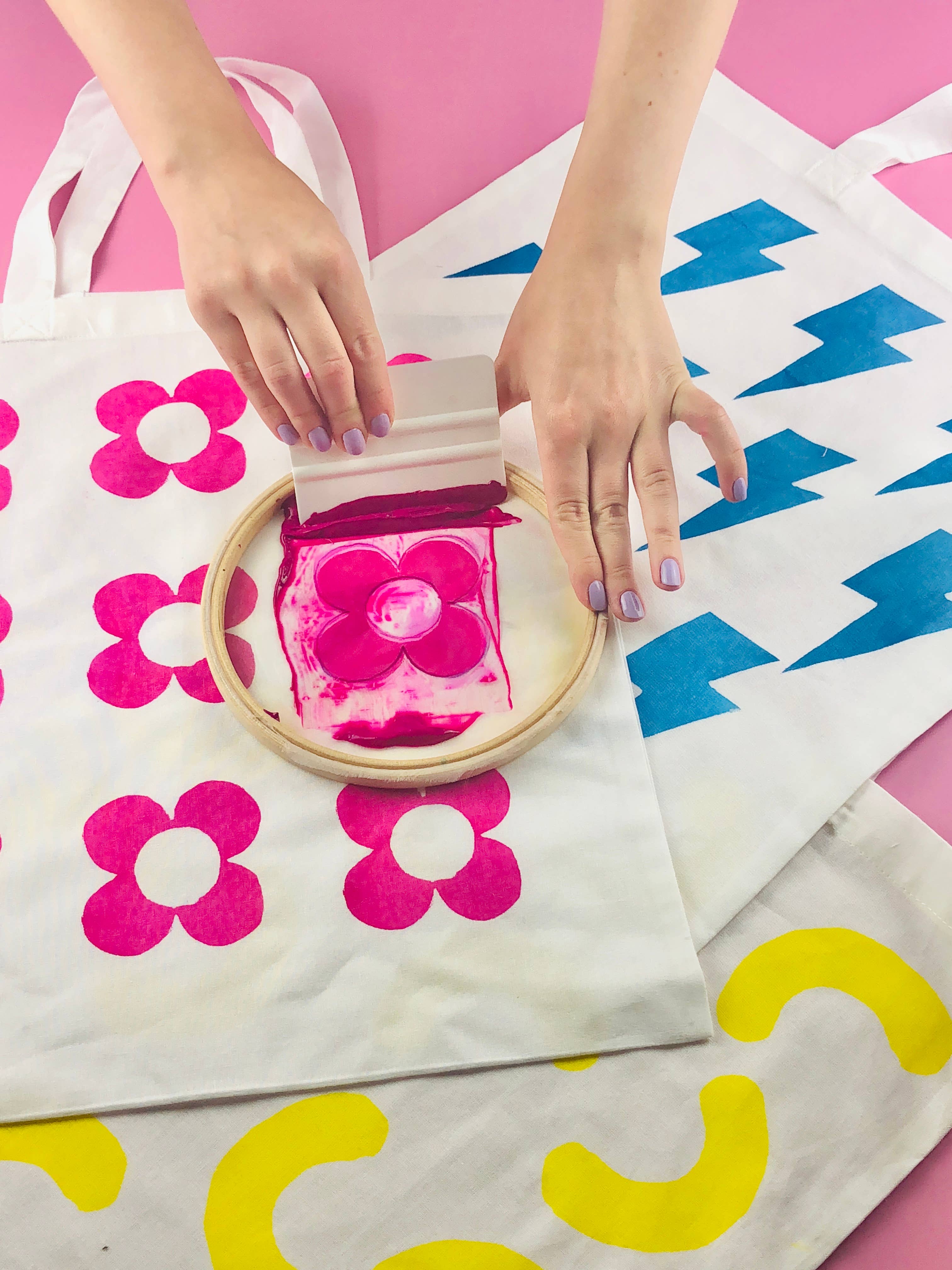 Person applying pink fabric paint to a white tote bag with floral designs on a pink background.