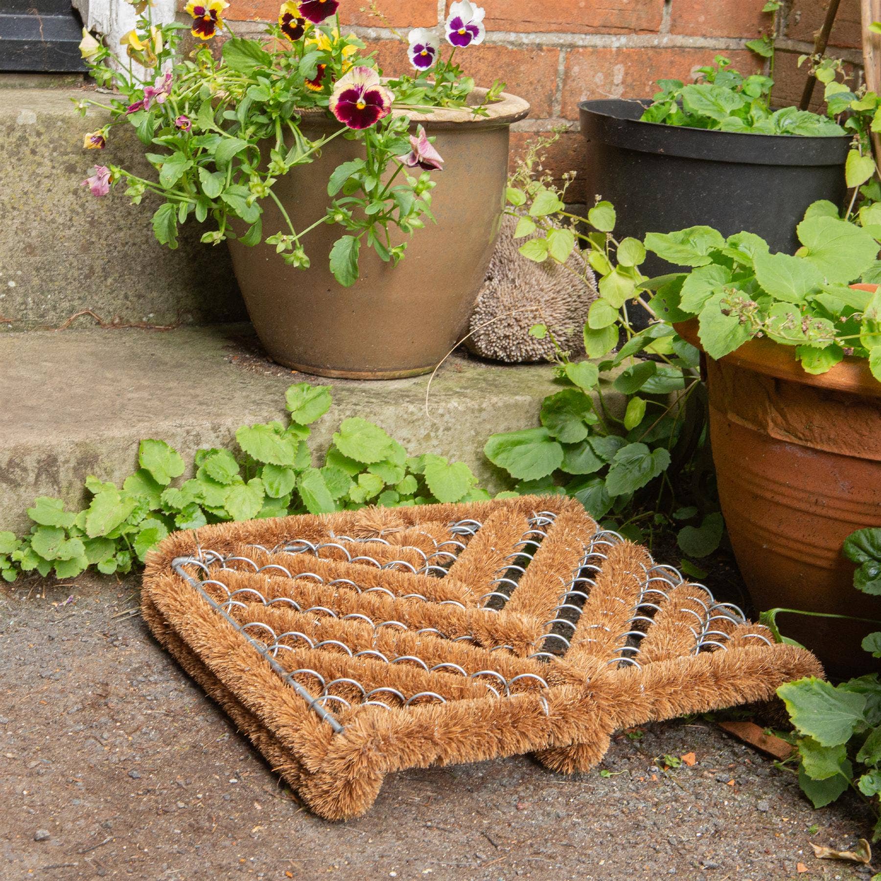 Brown doormat on a stone surface surrounded by potted plants.