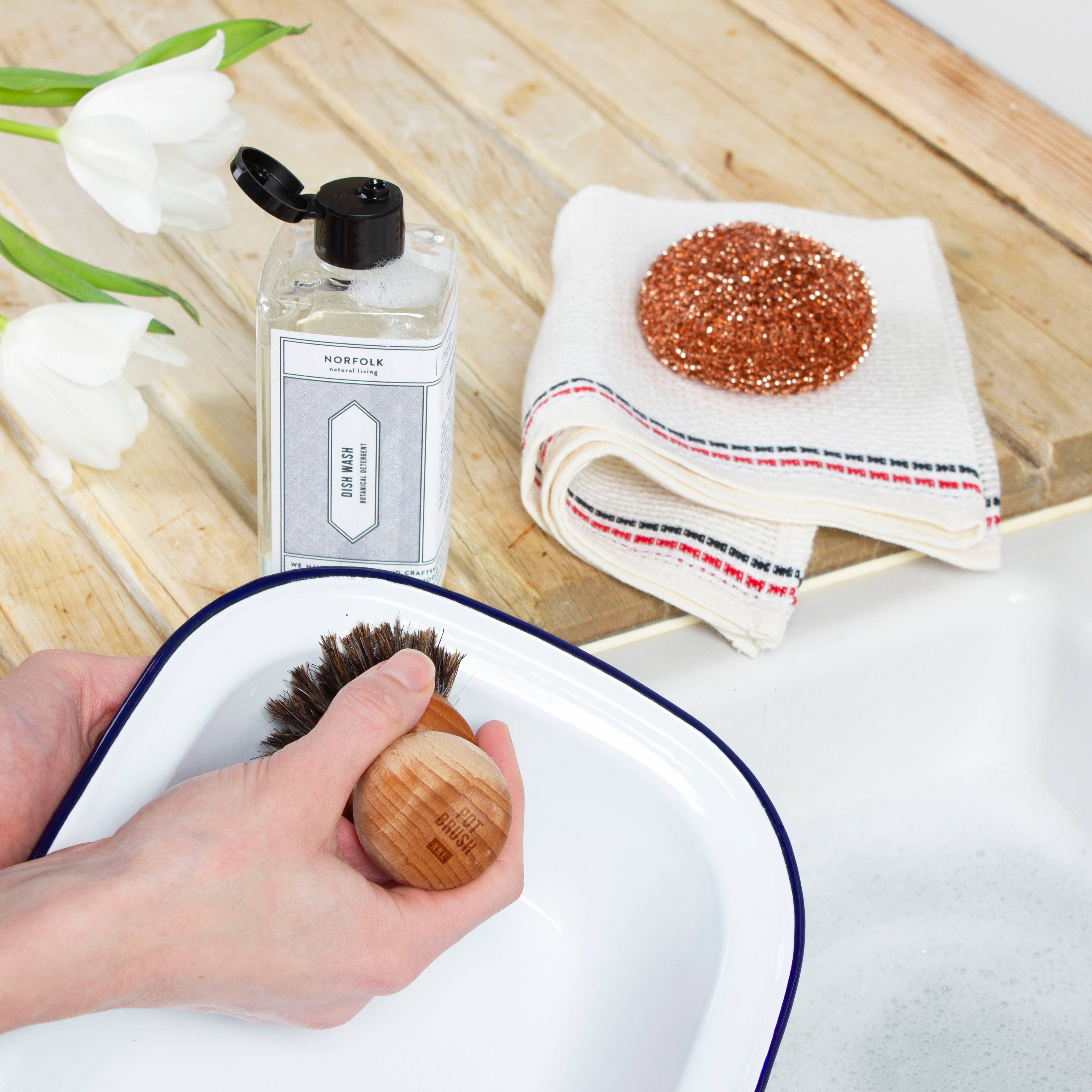 Person cleaning a dish with a brush, surrounded by a bottle of soap, a towel, and flowers on a wooden surface.
