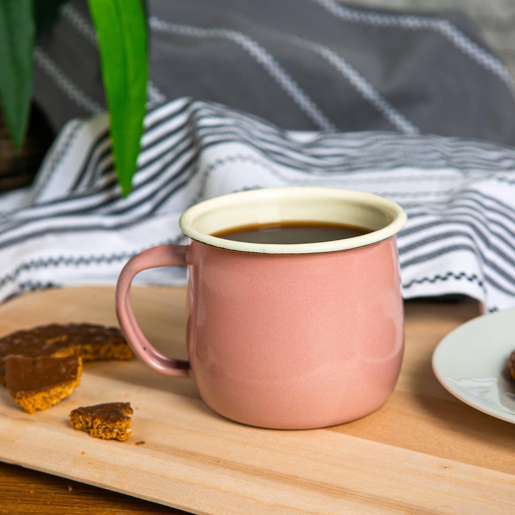 Pink mug with white interior on a wooden surface with cookies and a striped cloth in the background