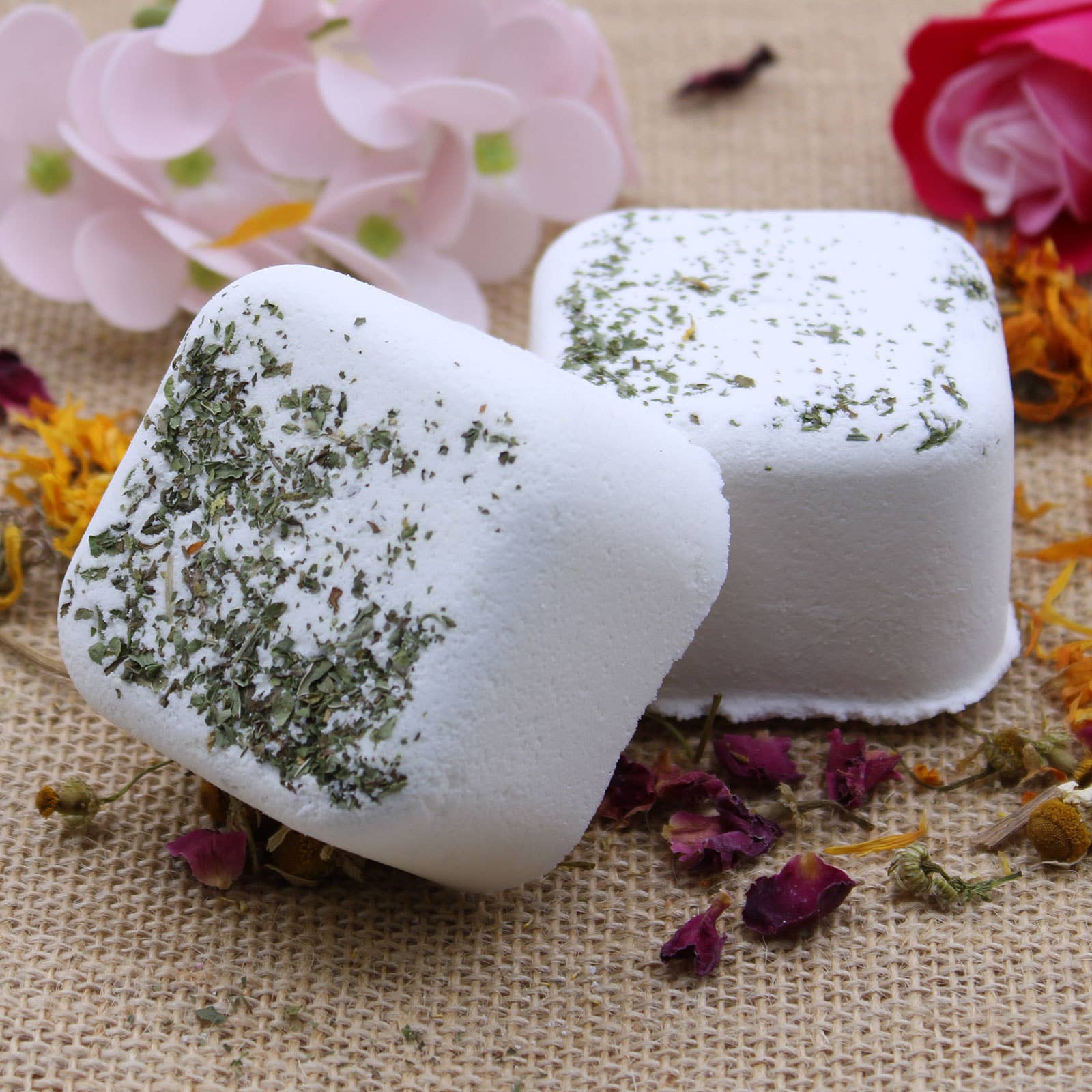 Two white bath additives with green herbs on a textured surface with flowers and petals.
