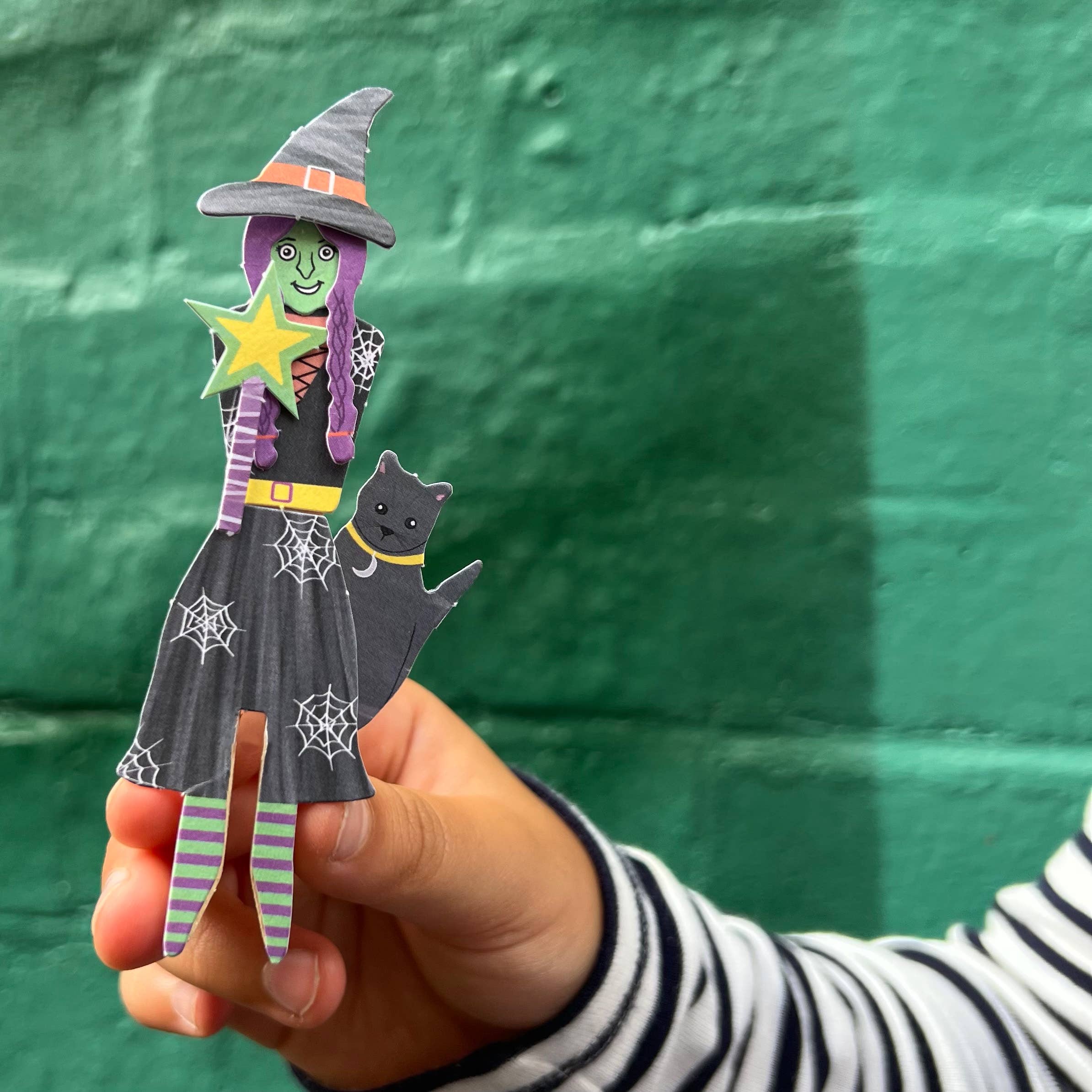 Hand holding a Halloween-themed paper cutout of a witch with a cat against a green wall.