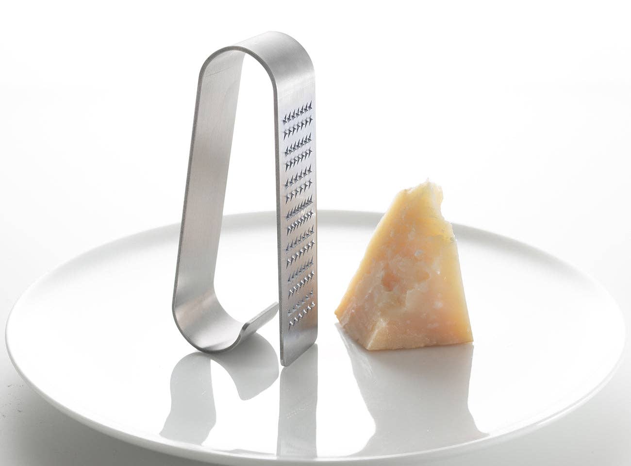 Metal cheese grater next to a wedge of Parmesan cheese on a white plate