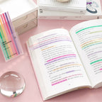 Open book with colorful highlights on a pink surface, next to a box of highlighters.