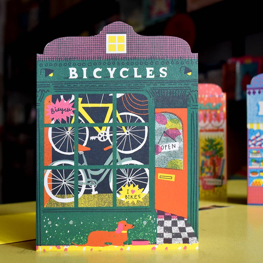 Colourful bicycle shop greetings card with a dog illustration on a yellow surface.