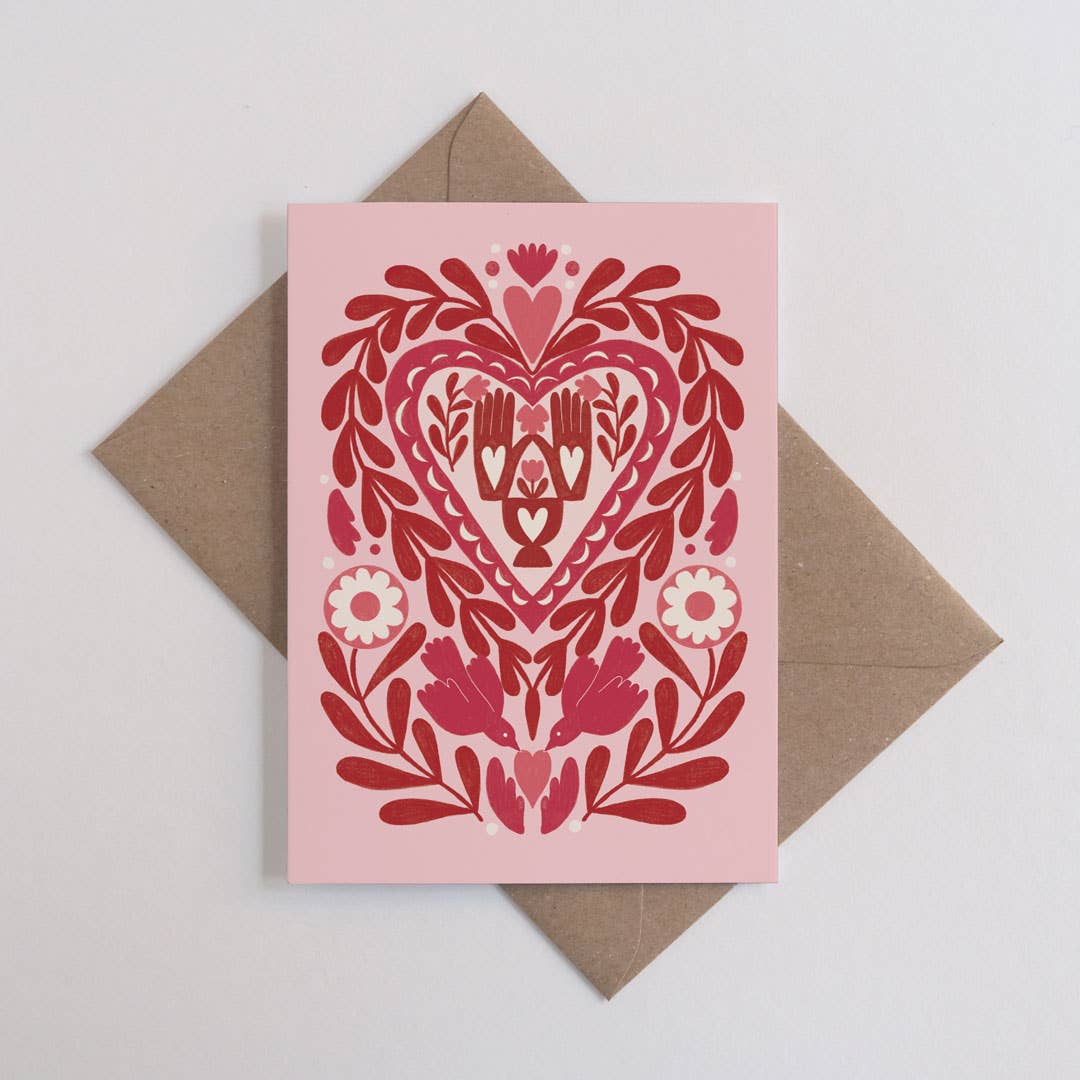 Card with red floral and heart design on pink background, placed on a brown envelope.