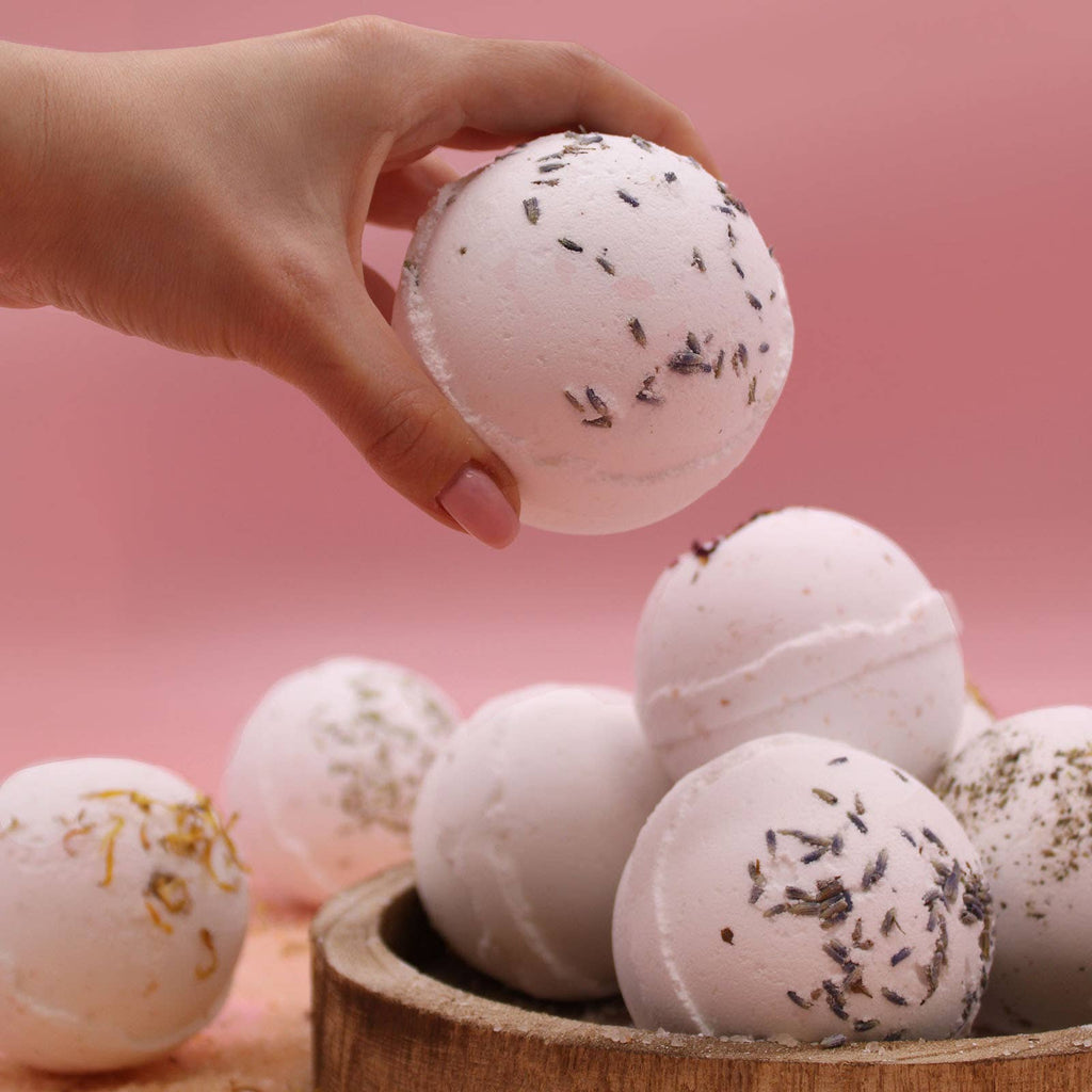 Hand holding a bath bomb with more bath bombs in a wooden bowl on a pink background