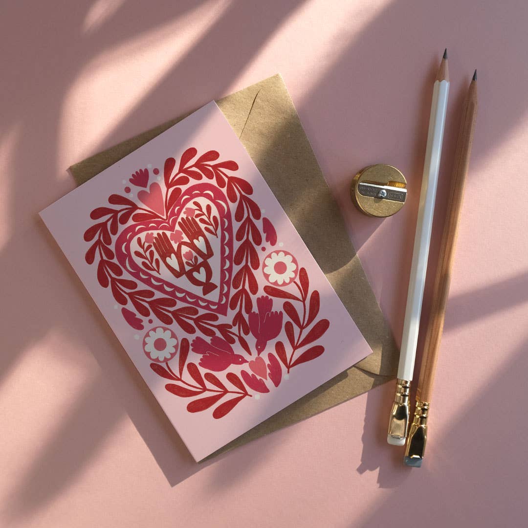 Greeting card with red floral heart design on a pink surface with a pencil and sharpener.