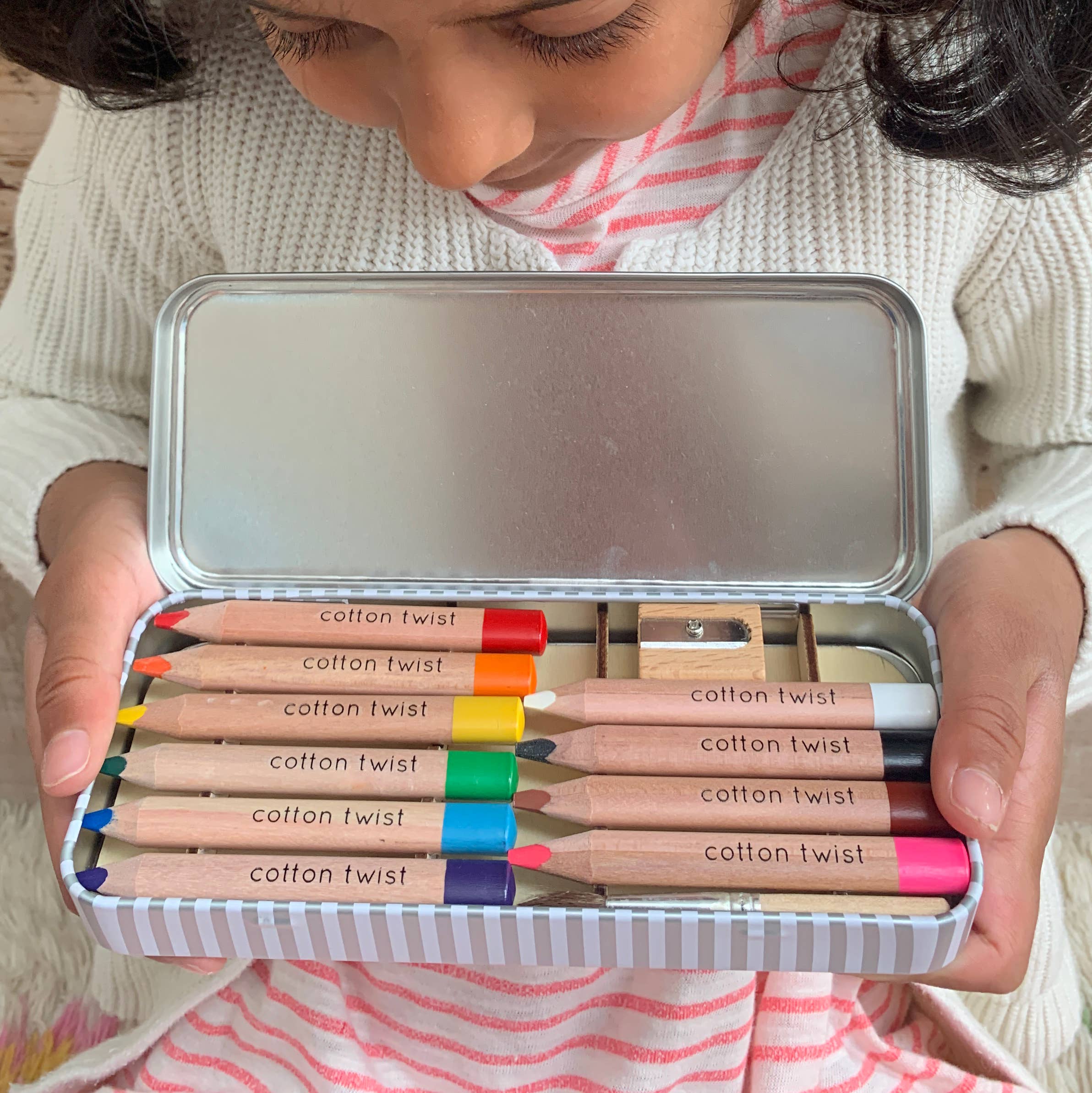 Child holding a set of Cotton Twist colored pencils in a metal tin.
