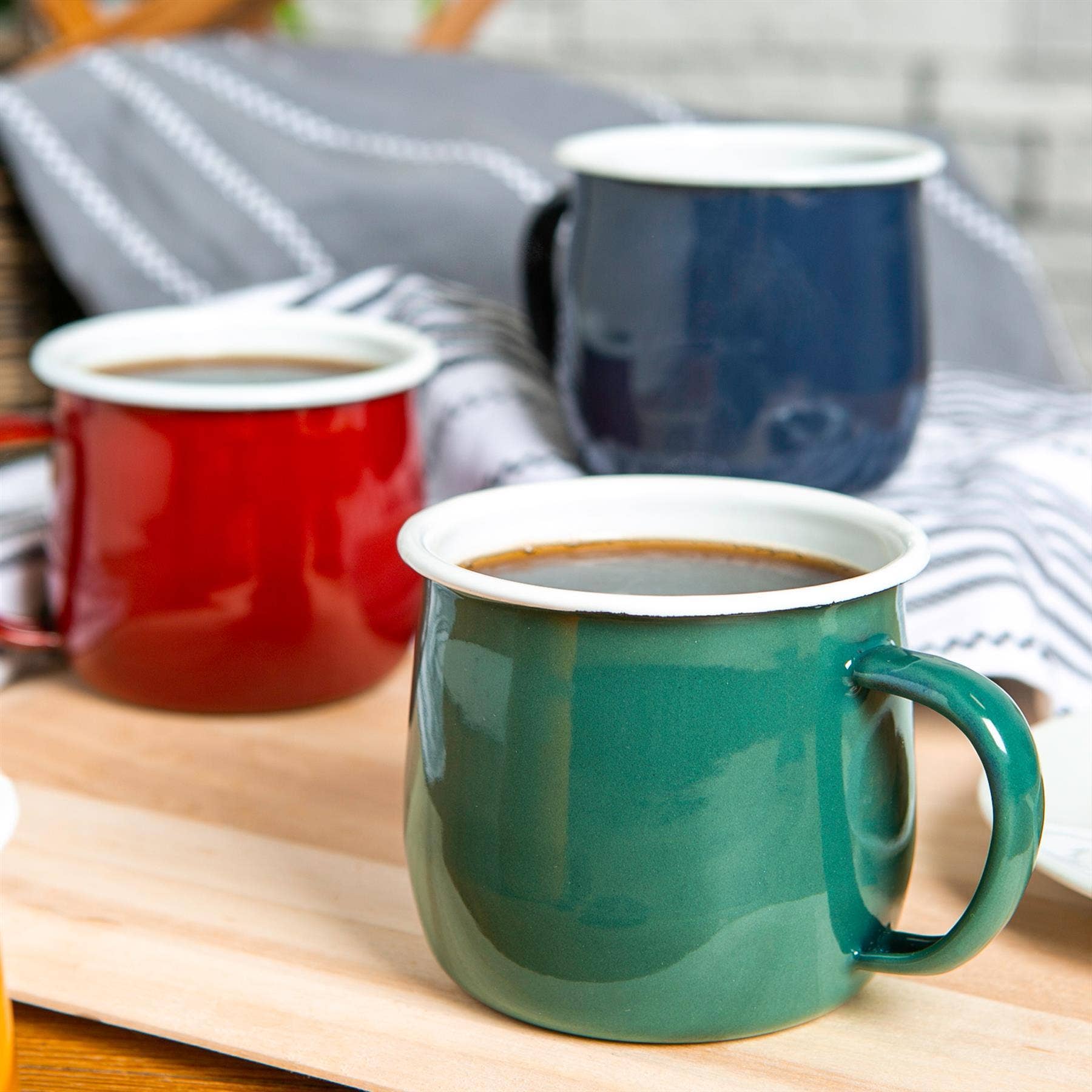 Three mugs in red, blue, and green on a wooden surface with a striped fabric background.