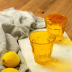 Two yellow glass cups on a wooden surface with a gray cloth and lemons.