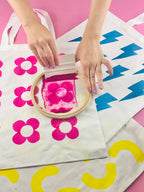 Person applying pink fabric paint to a white tote bag with floral designs on a pink background.