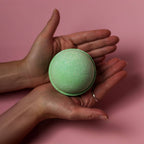 Green bath bomb held between two hands against a pink background