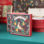 A square Christmas match box with a red border and an illustrated bird perched among pears and berries, with the text 'MATCH THE MOMENT?' and 'MY TRUE LOVE GAVE TO ME'. It sits on a teal surface with other holiday items blurred in the background.