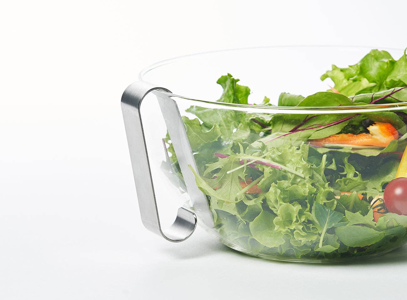 Clear glass salad bowl with metal handle filled with green salad on a white background