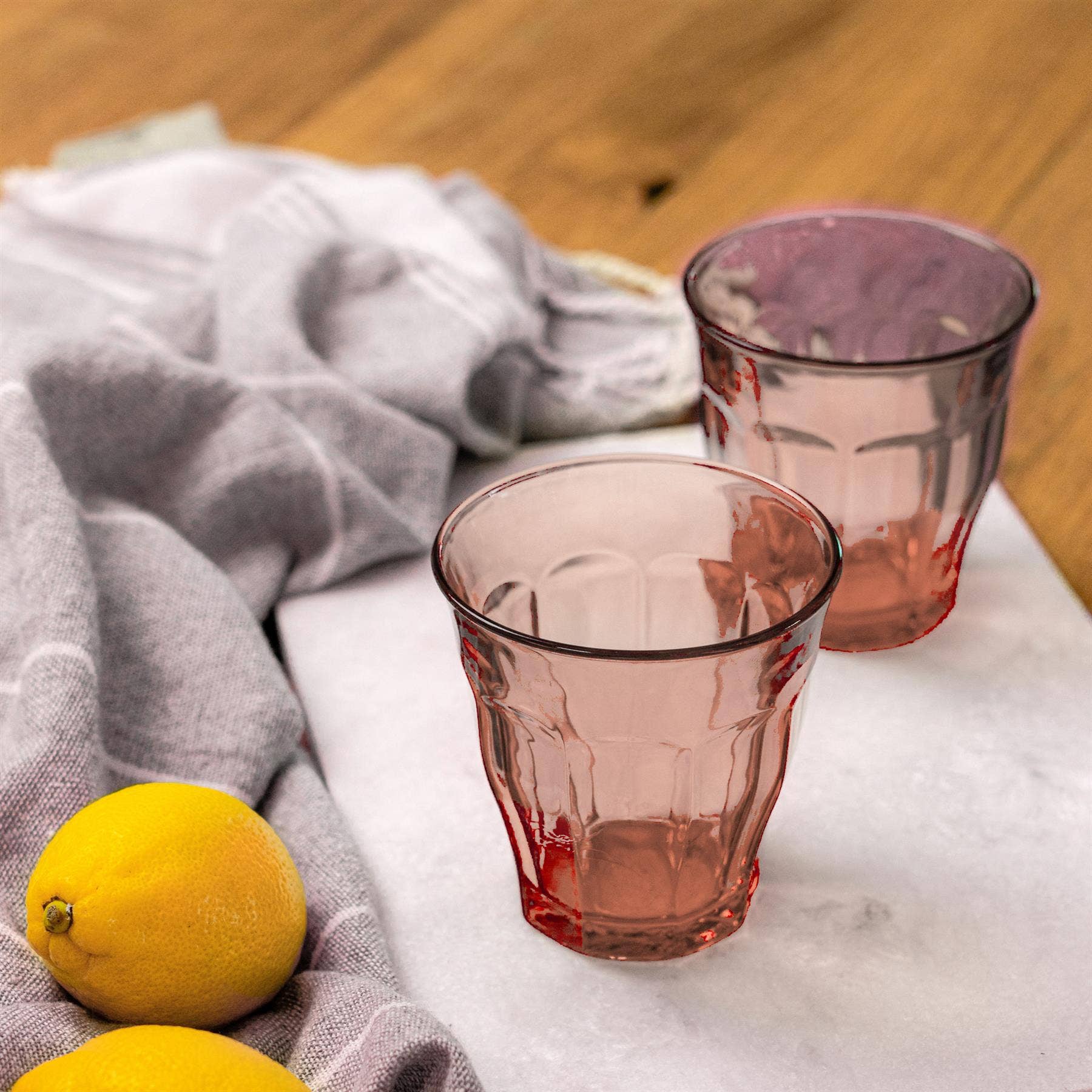 Two pink glass tumblers on a marble surface with lemons and a gray cloth.