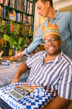 Man wearing a birthday hat sitting at a table with a cake, surrounded by books and plants.