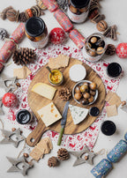 Assorted cheeses, crackers, and small dishes on a wooden board with festive decorations and drinks.