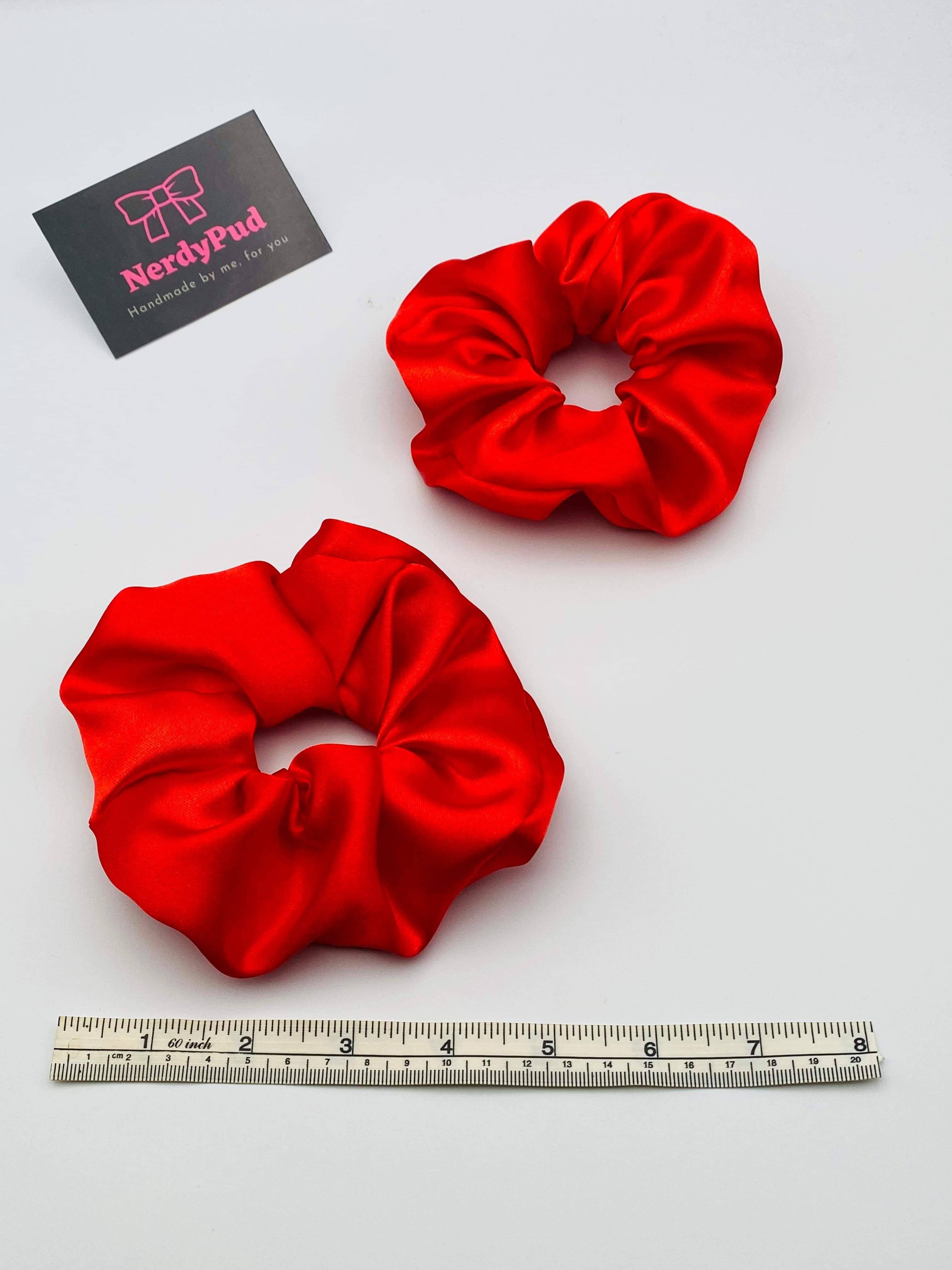 Two red scrunchies on a white background with a ruler for scale and a business card.