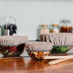 Three glass bowls with fabric lids on a wooden surface, containing various ingredients.
