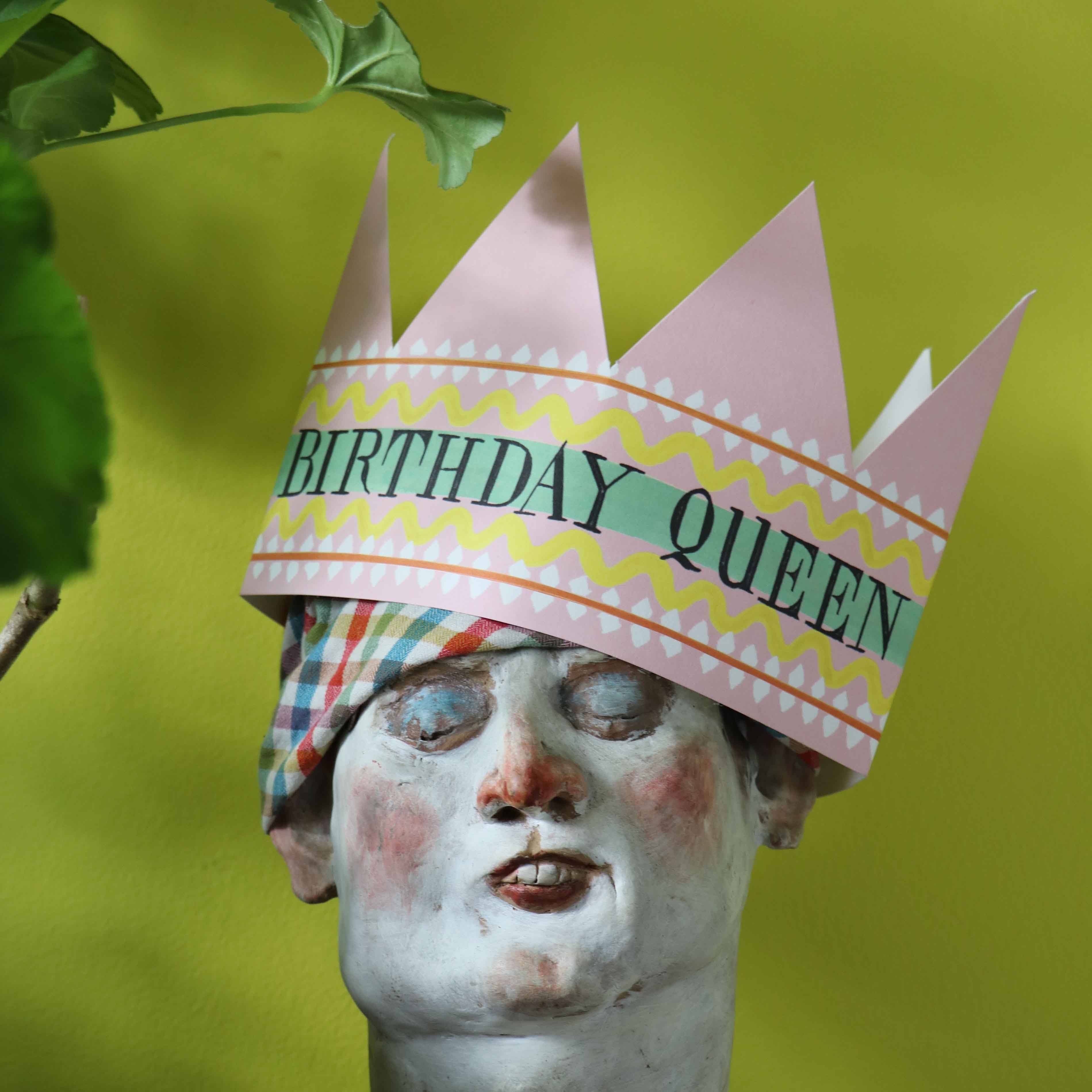 Statue head wearing a 'Birthday Queen' paper crown against a green background