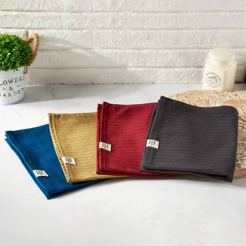 Four folded cloths in blue, yellow, red, and gray on a white surface with a textured wall background.