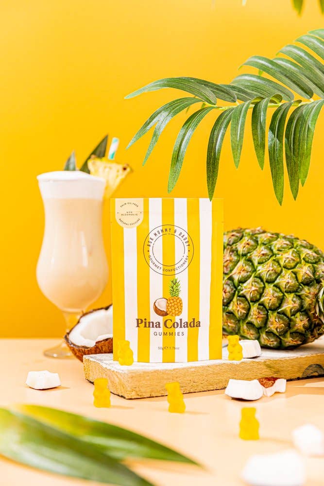 Striped bag of Pina Colada Gummies on a yellow background, styled with a cocktail glass and pineapple - All Good Things