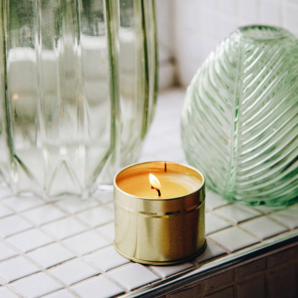 A lit silver tin candle showing light orange soy wax, set on white tiles with green glass vases in the background | Amber - All Good Things