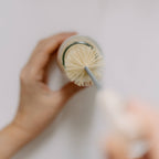 Top-down view of hands using a natural sisal brush to clean a glass bottle, against a white background - All Good Things
