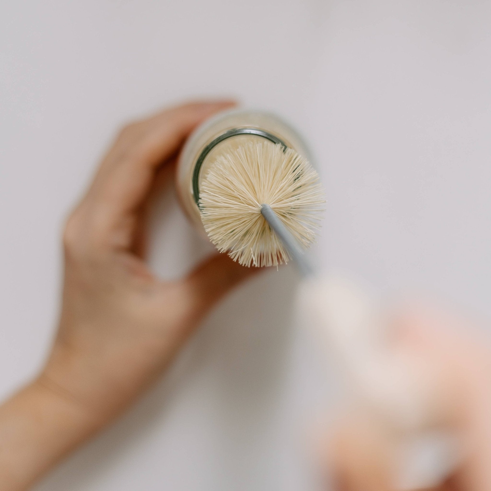 Top-down view of hands using a natural sisal brush to clean a glass bottle, against a white background - All Good Things