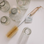 Top-down view of glass bottles on a white background with a natural sisal bottle brush lying alongside them - All Good Things